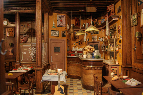 A detailed miniature model of a vintage cafe interior with wooden furniture, decorative bar counter, hanging lamps, and various accessories. Vintage posters adorn the walls, and there is a tiled floor.