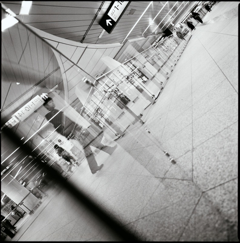 black and white square photograph of a series of reflections in the shibuya metro station. a glass barrier recedes into the frame, a man glances back and down at the lens.