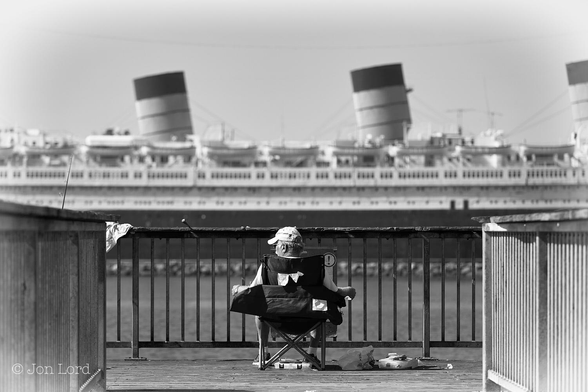 This is a black and white street photo in landscape format of a man fishing at the end of a small pier with a large ship in the background. Long Beach, California (2016).

Stretching away from the camera along the pier on both sides of the image are two sets of metal railings that appear to converge slightly with increasing distance. At the end of the pier and about five metres away is our fisherman, sitting in a dark coloured, fabric covered, picnic chair. His head is just visible above the back of the chair, he is looking to his left, as if aware someone is watching / photographing him. He is wearing a white baseball cap and dark sunglasses. His arms are lying on the arm rests at the sides of the chair. His two legs, just visible, with white socks and shoes. Under the chair and to the right is a jumble of ....stuff ? perhaps something to do with fishing ? A fishing Rod (fishing pole) is visible two metres to his left, resting against the far railings. Beyond the end of the pier is a body of calm medium grey water with a small stone breakwater at the far side. Next to the breakwater is a moored, large ocean liner, slightly blurred out of focus. The hull is black and lined with small portholes. The upper part of the hull and superstructure above, is white, this time lined with square or rectangular windows. Above the upper deck is a row of nine open lifeboats hanging from davits. There are three large, raked funnels beyond the lifeboats in the centre of the ship. 