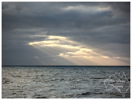 A dramatic landscape photograph taken on the Bunbury coast, showing a vast, dark, textured ocean filling the lower two thirds of the frame.  Above, the sky is heavy with dark grey, overcast clouds. A central break in the clouds allows bright, warm sunlight to stream down in distinct rays (crepuscular rays) onto the water and horizon.