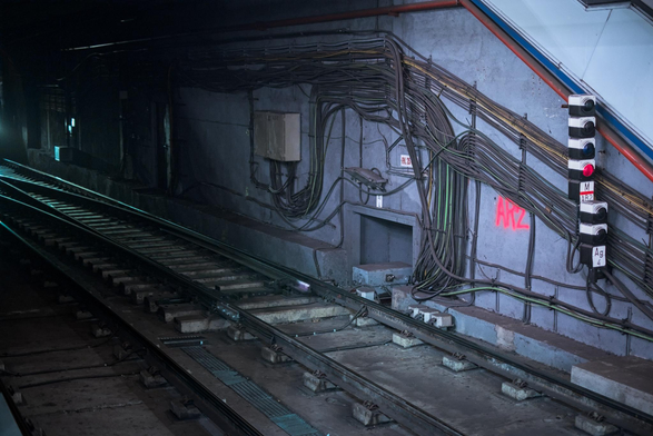 Fotografía urbana de un detalle en un tunel del metro. Cruzando el cuadro en diagonal se ven las vias del metro, iluminadas por las luces de un tren que no ha entrado en el cuadro. El muro del tunel en frente esta repleto de cables de distintos colores quie van en diferentes direcciones, un grupo de luces de señalización estan a la derecha del cuadro. En spray rojo intento esta la leyenda AR2 que destaca por el contraste de color.