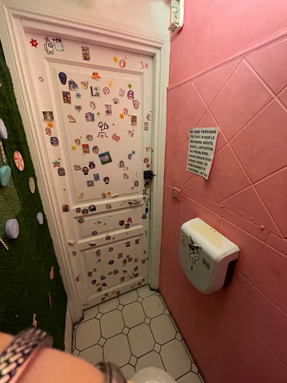 A cozy tiny room with a framed door partly covered with tiny LGBT and cute stickers. One wall to the right is pink, the other to the left is covered with fake green grass.
