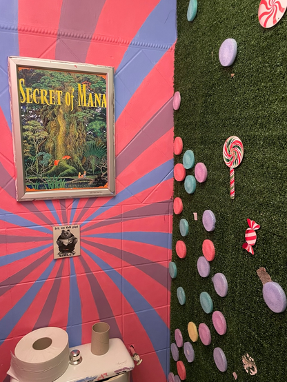 The fake green grass is plastered with lollipops, candies and various round colored ornaments. The last wall is painted with a bright pink, blue and lavender pattern and features a Secret of Mana poster.