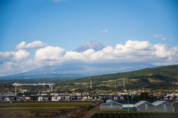 Mount Fuji seen in the distance, circled by scattered puffy white clouds. There’s rolling hills in the distance towards the mountain, covered in green trees and fields. In the near distance there’s a village of two-storey buildings farms, and many power line poles, pus a few viaducts bridging the hills nearby. The sky is a faint baby blue above it all.