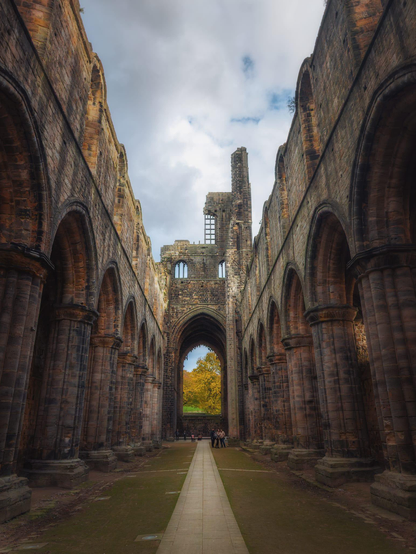 A dramatic, low-angle shot looking down the ruinous nave of Kirkstall Abbey in Leeds. Two rows of towering, weathered stone arches and pillars frame a central pathway that leads towards the main archway of the chancel, the roof and vaulting being entirely absent. Through the central arch, a partially collapsed tower and further ruins are visible, with a burst of brilliant golden-yellow autumn foliage from a tree in the distance contrasting against the muted brown and grey stone. A small group of people are gathered near the chancel archway, giving a sense of scale to the massive structure. The sky above is a mix of bright white and grey clouds.
