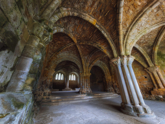 An interior view of the Chapter House at Kirkstall Abbey, showcasing its intricate medieval architecture with ribbed, vaulted stone ceilings supported by heavy columns. The space is dimly lit, with natural light entering through two small, high arched windows at the far end, illuminating a set of stone steps and a raised platform. The stone work is heavily weathered, displaying tones of mossy green, brown, and warm orange-red, creating a sense of age and ruin. The perspective is slightly wide-angled, emphasising the curve of the arches and the impressive height of the central pillars.
