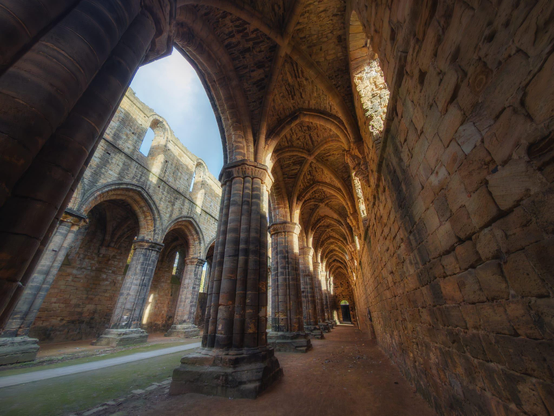 A dramatic, low-angle photograph captures the interior of the ruined nave and aisle of Kirkstall Abbey, showing the difference in preservation between the two sections. On the left, the main columns of the nave stand mostly intact, supporting the remains of the arcade arches, with the sky visible through the missing roof. On the right, the aisle is relatively complete, featuring an impressive perspective of a long, vaulted stone ceiling supported by a continuous row of columns, leading into the distance. Bright sunlight streams through the open side of the nave, contrasting with the shadowed aisle, where the rough, textured stone walls and ceiling are illuminated with a warm, golden-orange light.
