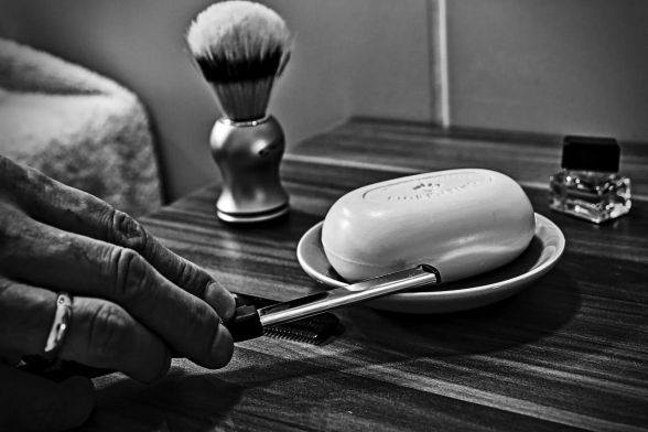 Auf einem kleinen Badezimmerschränlchen stehen ein Rasierpinsel, ein Eau de Toilet und eine kleine Schale mit Rasierseife. Eine Hand streift das alte Rasiermesser an der Schale ab. 