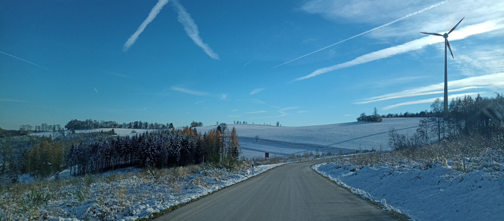 Aufnahme aus dem fahrenden Auto, das graue Straßenband wird eingerahmt von einer hügeligen Schneelandschaft überspannt von einem blauen Himmel