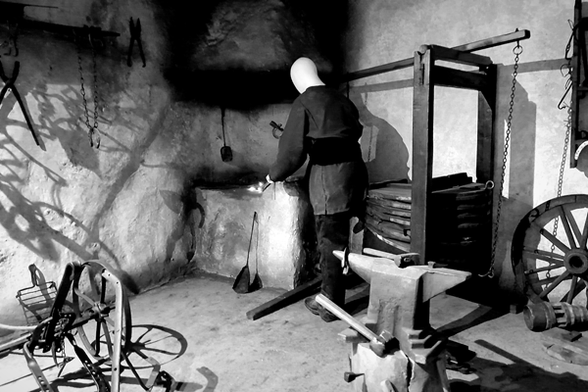 The picture is black and white. It shows the inside of a forge focusing on the corner of the room. The sides of the pictures are brightly lit creating sharp shadows, the corner that's in the middle of the image is more shadowed. The head of the featurless blacksmith mannequin contrasts sharply with its dark background. He is turned with the back to he viewer, forever working on its nonexistent project.