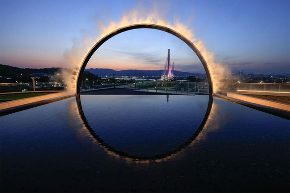 A stunning view of a circular arch glowing with a smoke effect, reflecting in still water, with a bridge lit in purple against a twilight sky.