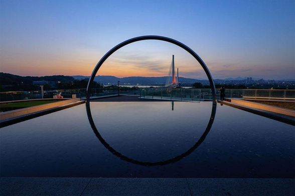 A stunning view of a circular arch reflecting in still water, framing a bridge lit in orange in the distance against a twilight sky.