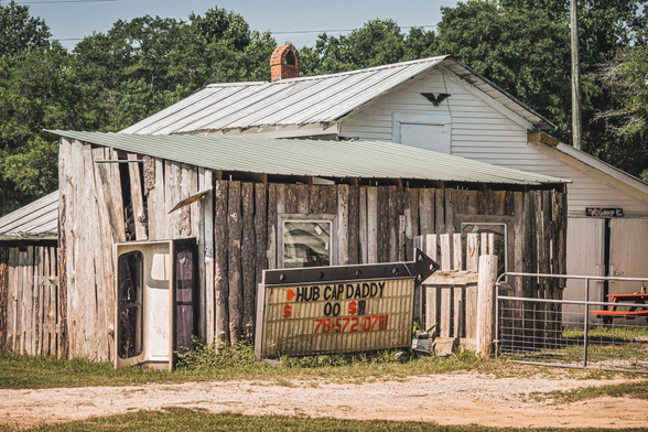 Dilapidated rural roadside building with severely weathered vertical wood siding showing extensive deterioration and missing sections. The structure has a corrugated metal roof in poor condition. A rusty directional arrow sign reading 'HUB CAP DADDY' with a phone number '79-572-07' is mounted on the front. Behind it stands a white wooden house with horizontal siding, a metal roof, and a red brick chimney. Dense green trees fill the background under an overcast sky. A metal gate is visible on the right side, and the foreground shows a dirt and gravel area with patches of grass.