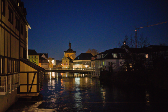 Den Fluss entlang fotografiert. Das Bamberger Rathaus ist zu erahnen in der Mitte. Baidseitig Häuser. Altstadt. Dunkel Beleuchtug. Ein Kran Überragt die Dächer.