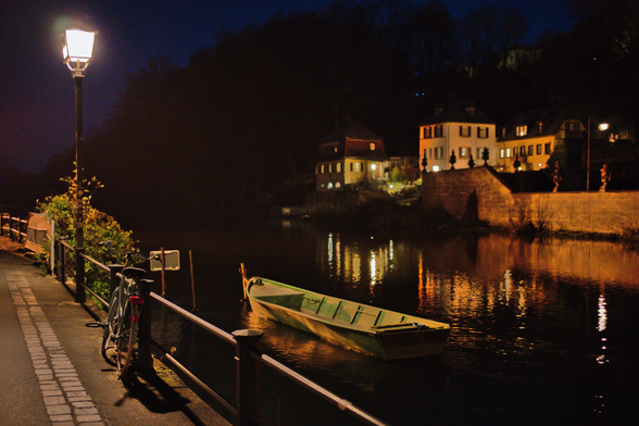 Am Fluss entlang fotografiert. Ein Holzboot ist im Fluss. Eine Lampe am Ufer. Daran ein Fahrrad. Dunkel. Die Lampe leuchtet. Auf der anderen Seite des Flusses Beleuchtete Häuser. Es geht in Wald über.