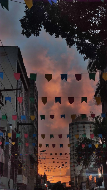 O céu alaranjado de fim de tarde contrasta com os prédios e árvores. Bandeirinhas coloridas de são João decoram a rua, criando um clima festivo.