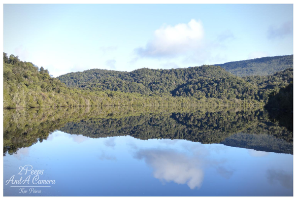 Colour landscape photograph of the Gordon River in Tasmania, signed by Kev Peirce Photography.

The image features a wide expanse of still, dark water perfectly reflecting the dense, deep green temperate rainforest covering the steep hillsides that frame the river.

The reflection is almost a perfect mirror image, including the small patches of white cumulus clouds in the pale blue sky above. The scene captures the tranquility and pristine nature of the remote wilderness.