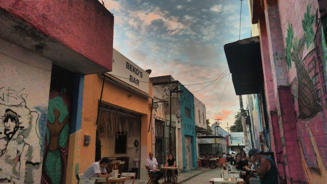 Uma rua vibrante com prédios coloridos e grafites. Um bar, "Beko's Bar", é visível. Pessoas sentadas em mesas ao ar livre desfrutam do final de tarde, sob um céu com nuvens alaranjadas.