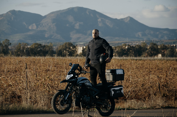 Rider standing beside a dual-sport motorcycle equipped with panniers and top box on a rural paved road, with dry fields and distant mountains under a clear sky.