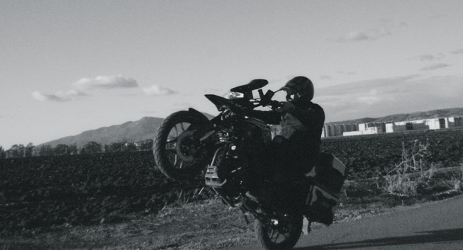 Black-and-white image of a rider performing a wheelie on a dual-sport motorcycle along the same rural road, with buildings and mountains in the background.