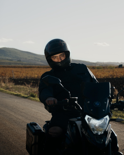 Rider in black gear and helmet seated on a black BMW motorcycle on a rural road, facing the camera with headlight on and hills in the background.