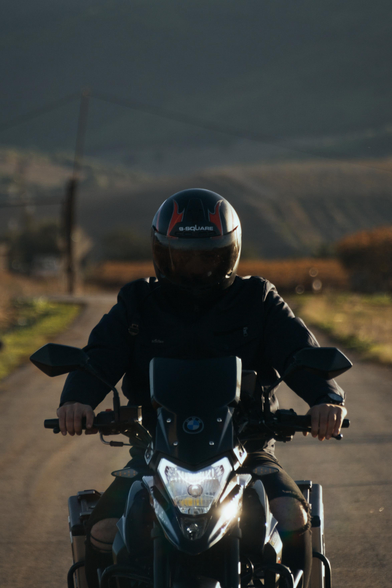 Rider wearing a black helmet with red and white accents, riding a BMW motorcycle on a narrow countryside road with hills and power lines in the background.