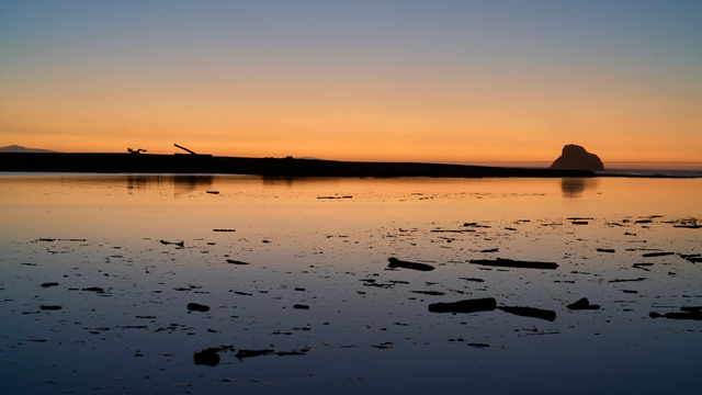 The fading light of day after the sun goes down. The light is orange and reflected in the river. There is a sand bar across the horizon with a couple pieces of driftwood. There is a sea stack just beyond. Floating in the foreground is debris dislodged by the rising tide.