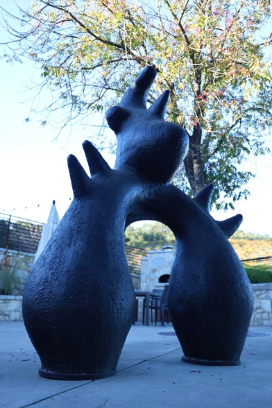 A large, abstract black sculpture stands outdoors, featuring organic shapes with a central arch. In the background, a tree with green leaves and a stone wall is visible, along with outdoor furniture.
