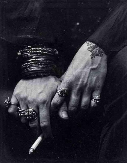 Close-up black and white photograph of two hands adorned with multiple ornate rings and bracelets, one hand holding a cigarette.