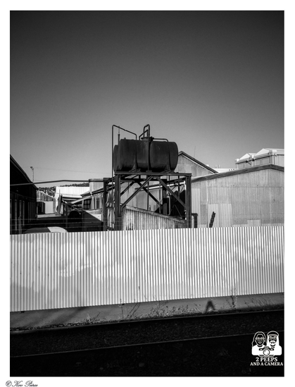 A striking black and white photo of an industrial scene. In the foreground, a low corrugated iron fence runs horizontally across the frame. Behind it, on a black metal frame, sit two large, rusty, cylindrical tanks.

Beyond the tanks are various corrugated iron sheds and industrial buildings, all under a vast, bright sky. A single railway track runs along the bottom of the image, below the fence line.