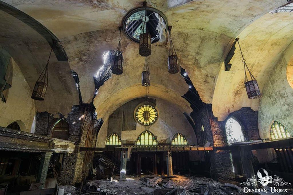 Interior of an abandoned building with arched ceilings, broken windows, hanging light fixtures, and debris covering the floor.