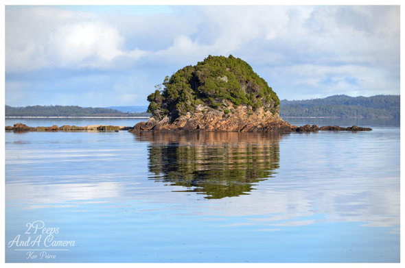 A tranquil, wide shot photograph of a small, dome shaped rocky island covered in dense green scrub and trees, resting in the smooth, mirror like blue waters of Macquarie Harbour.

The island's reflection is perfectly cast on the surface of the water, and the distant shore, covered in forest, sits under a bright blue sky with scattered white clouds. Photo by Kev Peirce.