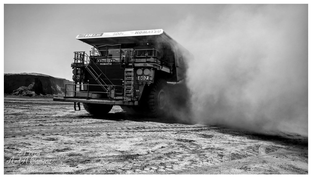 A dynamic, low angle, black and white photograph signed by Kev Peirce. A massive Komatsu 830E-AC haul truck, identified by its model number and '802' on the side, is seen from the rear quarter.

The truck is driving on a dusty, cracked dirt surface, generating a huge, thick cloud of dust that billows dramatically to the right side of the frame.

In the background, there is a large, sheer embankment of excavated material. The image captures the scale and activity of a major mining operation.