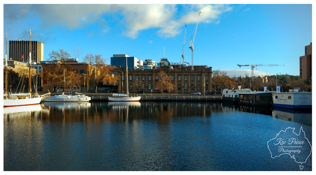 Colour photograph signed by Kev Peirce, capturing an early morning scene at the Hobart waterfront. Several sailboats and a covered white boat are moored in the dark blue water in the foreground.

Behind them, historic stone buildings with autumn coloured trees line the shore. Construction cranes rise above the buildings against a bright blue sky with white clouds.