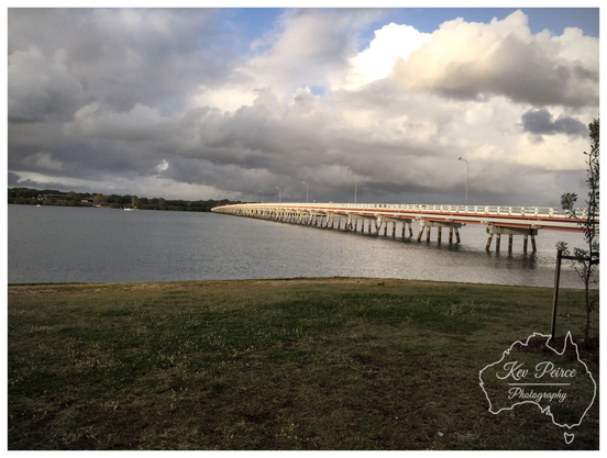 A wide angle landscape photograph of the Bribie Island Bridge spanning calm, dark water under a cloudy, dramatic sky.   The low lying bridge extends from the right side into the distance, supported by numerous wooden pylons. The bridge deck has white railings and a prominent red-orange stripe along the side.  The foreground features a strip of grassy, slightly uneven ground. The sky is dominated by heavy grey clouds, with patches of bright white and blue breaking through near the center.