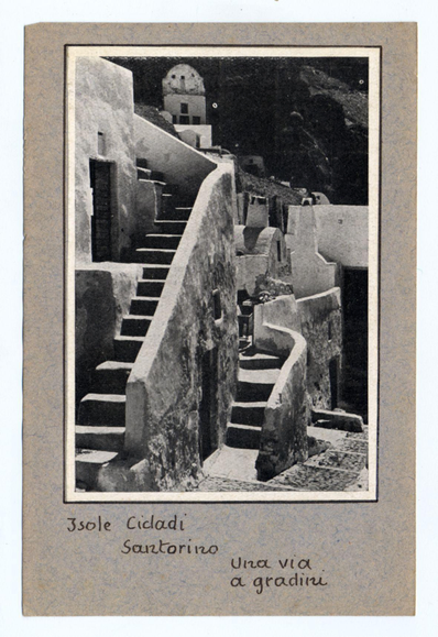 The image depicts an old, monochromatic photograph of winding steps leading up a steep hillside in Santorini. The architecture is reminiscent of Cycladic structures found on the Greek island, characterized by its white-painted walls and simple design. A church with a dome is visible at the top of the stairs, suggesting that this could be part of a religious or historical site within the town. Text overlays indicate it's an old postcard from Santorini featuring "3sole Cialadi" (three suns) as mentioned on one side and "Una via a gradini" which translates to 'a way with steps' on the other, indicating these may be used for climbing or walking up slopes. The image appears aged with some wear marks visible around its edges, adding to its vintage charm.