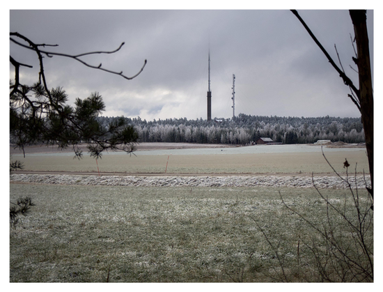 En liggande bild på två master i skogen längst bort. Den längsta masten sticker upp i de grå molnen. Träden runt om är varierande gröna och vita. I mitten några frostiga fält med en korsande väg. Förgrunden består av träd och grenar som ramar in.