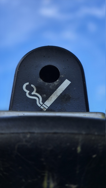 Detailaufnahmen eines öffentlichen Aschenbechers mit Zigarettensymbol vor blauem Himmel. 
