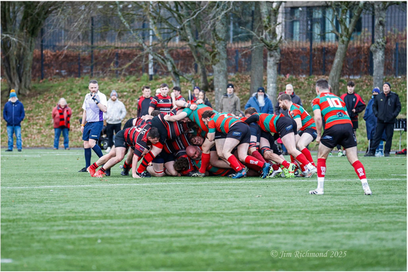 A rugby match scene with players in a collapsed scrum. The team in red and black stripes is engaged with a team in green and red stripes. A referee oversees the action, and spectators are lined up in the background. 
