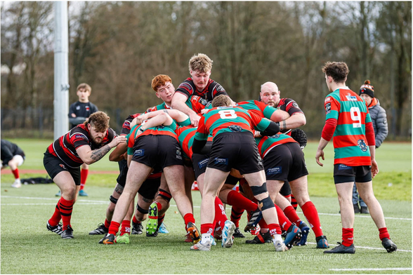 A group of rugby players is engaged in a scrum on a grassy field. Players wear red and green jerseys, and there are additional individuals in the background preparing for the game. Trees and a goalpost are visible in the surroundings.