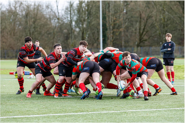 A rugby match in progress on a grass field. Players in red and black jerseys are engaged in a scrum, while one player in a green and red jersey prepares to pass the ball. Other players are observing the play.
