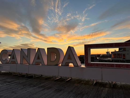 A large three-dimensional “CANADA” sign is backlit by a colourful sunrise on a wooden boardwalk. The dark brown boards run perpendicular to the sign, which is shiny and white with thin red outlines. To the right of the letters is a rectangular feature that frames a docked ferry. The sky has thin clouds emanating from the image center near the horizon, with the sky colours ranging from bright blue at the top to yellow to deep orange near the horizon. [Halifax (NS, Canada), November 2025]