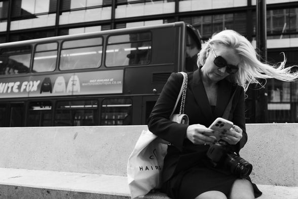 The image is a black and white street photograph featuring a woman seated on a concrete bench. The woman, with her head slightly bowed, is intensely focused on her phone, which she is holding in her hands. She has long, blonde hair that is being blown by the wind, creating a dynamic effect. She wears sunglasses, a dark blazer, and what appears to be a dress or skirt. A camera is resting on her lap. A small, quilted purse with a chain strap hangs from her shoulder, and a tote bag, with the word "HAIRS" printed on it, rests beside her.