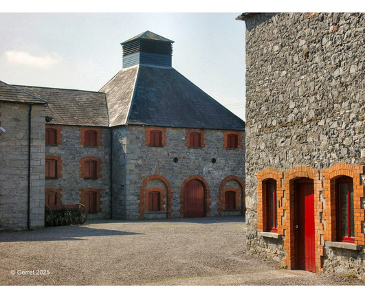 Historic stone distillery courtyard with arched red wooden doors and windows, lit by warm sunlight. The sky is clear, creating a serene atmosphere.