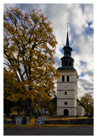 En stående bild på en vit kyrka med skriften ”CJU EEB 1741”, vilket är initialerna på grundarna av kyrkan och årtalet den stod klar. Till vänster en lövträd med orangea löv och marken är helt täckt med orangea löv. Himmelen molnig med blå öppningar.