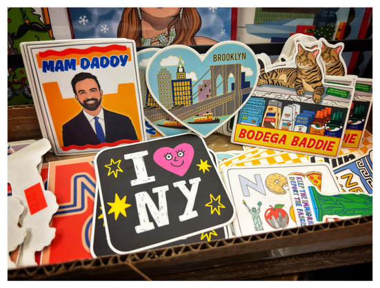 Photo of a collection of New York-themed stickers inside a cardboard box. Among the most notable: an illustration of a smiling bearded man in a suit and tie (Mayor-Elect Zohran Mamdani) under the text “Mam Daddy,” a heart-shaped sticker with a graphic of the Brooklyn Bridge, an illustrated cat lounging on a store shelf with the text “Bodega Baddie,” and a hand-drawn “I (Heart) NY” image with a smiley face on the heart.