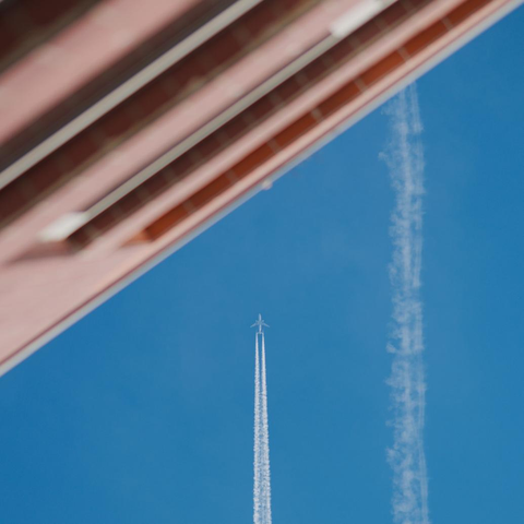Abstract photo: the top left trangle (one third of the image) is filled with a beige house that's out-of-focus. The other two thirds are filled with a blue sky. On the right of the sky is an old fuzzy plane trail. In the middle of the picture, coming vertically from the bottom, is a plane leaving a fresh trail.
