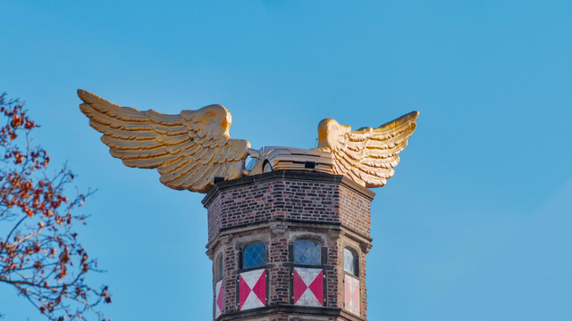 Foto eines vergoldeten Autos mit goldenen Flügeln auf einem Turm aus Backstein, vor blauem Himmel