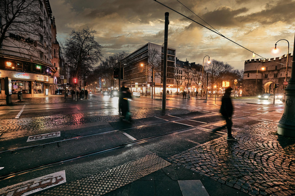 Wide-angle view of a large city square at twilight after a recent rain. Tram tracks and cobblestones in the foreground, glistening wet. A couple of passers-by as slightly motion-blurred silhouette figures. The lights of the city glow and reflect on the ground along with the fading light of late sunset.