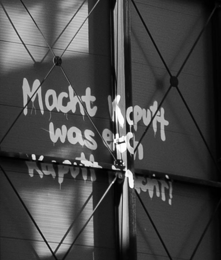 Motiv:
Graffito auf einer Wandfläche. Der Schriftzug lautet „Macht kaputt was euch kaputt macht!“. Metallstreben verlaufen kreuzweise über die Fläche. Schatten und Reflexionen liegen auf der Oberfläche.

Stimmung & Wirkung:
Kontraste durch harte Schatten. Betonung der Struktur durch Licht und Linienführung. Verdichtung durch Überlagerung von Text und Metallstreben.

Technische Details:
Bildformat: Street-Photography / Dokumentation.
Farbe: Schwarzweiß.
Schärfentiefe: Durchgehend scharf mit klar erkennbaren Strukturen im Vorder- und Mittelbereich.
Lichtverhältnisse: Direktes Licht mit starken Schattenwürfen und ausgeprägten Reflexionen.